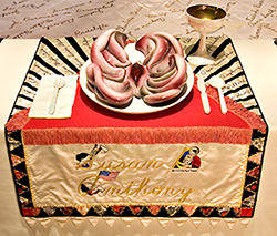 Foto av en tallrik och bestick på en broderad duk med Susan B. Anthonys namn på. En dricksskål i guld står också på bordet