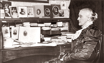 Foto i halvfigur av Susan B. Anthony sittande i profil framför sitt skrivborde, som på två rader är fullt av foton på andra rösträttskvinnor