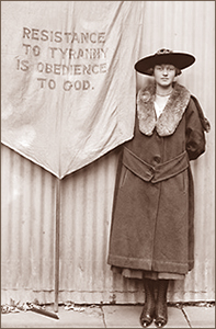 Foto av en kvinna i helfigur iförd hatt och kappa, och som står vid entt standar med texten "Resistance to Tyrrany is Obedience to God"