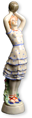 Porslinsfigur av tjej i blåprickig volangkjol som står med armarna över huvudet där hon håller i en boll, beredd att kasta