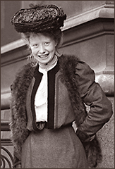 Foto av Mary Macarthur i halvfigur utanför en stenbyggnad. Hon har hatt och står leende mot kameran med ena armen i sidan