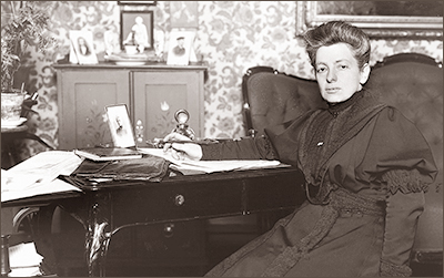 Foto i halvfidur av Gerda Meyerson som sitter vid ett skrivbord och ser in i kameran. Hon har en tjusig mör klänning. I bakrgunden syns en byrå med fotografier och en piedestal med en blomma