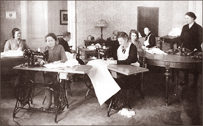 Foto av ett antal kvinnor som sitter eller står vid olika bord och symaskiner i ett rum