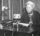 Foto av äldre Kerstin Hesselgren som sitter vid ett skrivbord och talar in ett föredrag för radio. Hon har papper framför sig och ser allvarlig ut.