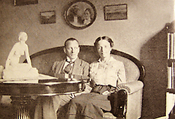 Foto av Albert och Elise som unga, sittande i en soffa. På väggen bakom dem hänger fotografier. På bordet framför dem står en statyett, kanske "Grodan". Vid sidan av Elise skymtar en bokhylla.