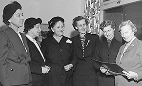 Foto av fem damer från Svensk damtidning samt Ottar som tittar på papper hon har i händerna. Alla i halvfigur och klädda i dräkt. De tre damerna till vänster har också hatt.