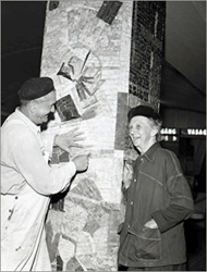 Foto av en man som arbetar med stenmosaikpelaren och Vera Nilsson som står och pratar med honom