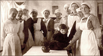 Foto av tio kvinnor som står runt ett bord, de har hucklen och förkläden på sig. På bordets ena sida sitter en liten flicka med rosett i håret