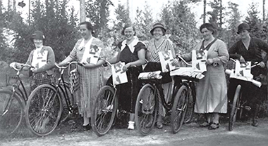 Foto av sex kvinnor som står med sina cyklar och massor av tidningar ute på en skogsväg. Alla kvinnorna ler eller skrattar mot fotografen