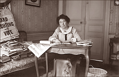 Foto av Christabel Pankhurst som sitter vid ett skrivbord mitt i bilden. Tidningen The Suffragette hänger ner från bordet och en papperskorg står bredvid. I bakgrunden syns dörrarna och till vänster en soffa belamrad av tidningar och böcker