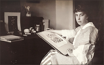 Foto av kvinna som sitter vid ett skrivbord med en ritbräda och på den en illustration. Hon ser in i kameran med allvarlig min.