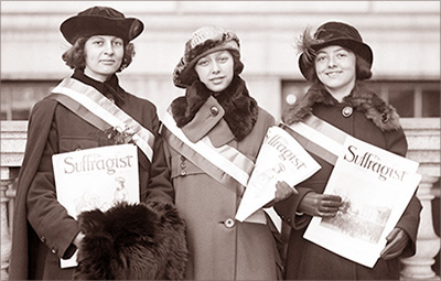Foto av tre unga kvinnor i halvfigur som står med kappor och hattar på gatan och håller i tidningar The Suffragist. De har också band över brösten med cu:s färger gult, vitt och lila, även om det inte syns på en bruntonad bild 