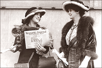 Foto av två kvinnor i halvfigur som ser på varandra och skrattar. Bäffe har hattar, den ena håller upp ett ex av Womans Journal samtidigt som hon håller en drös tidningar på samma arm, hon sträcker ut den andra armen och har en tidning i den handen