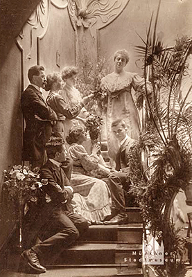 Tre män i kostym och fyra damer i tjusiga klänningar är draperade med blommor i trappan i Hovateljé Elvira.