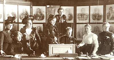 Foto av åtta kvinnor som sitter eller står vid ett bord. I bakgrunden syns stora porträtt av vad som mest verkar vara herrar