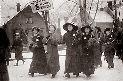 Foto av kvinnor i demontsrationståg. En bär ett plakat där det står "8 hour work day" på. Det är vinter, de är påklädda och det verkar snöa.