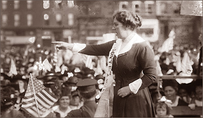 Foto av kvinna som står ovanför en folksamling och håller tal, hon pekar rakt ut med en armen. Nedanför syns kvinnor och barn och en vaktande polis. I bakgrunden syns husfasader.