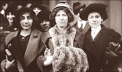 Foto i halvfigur av tre kvinnor som håller varandra under armarna, den till vänster har rufsigt hår under mössan och håller i en bok, den i mitten har pälskrage och pälsmuff, och den till höger har glasögon och håller i sin handväska. I bakgrunden anas fler folk, och en kvinna sticker fram huvudet mellan två av dem