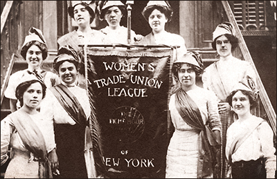 Foto av nio kvinnor som står i en trappa med konstiga luvor på sig, de håller i ett standar som det står "Women's Trade Union League of New York" på. De har också band över brösten, men utan text på. De flesta ser glada ut.