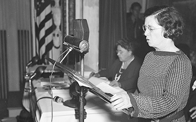 Foto i halvfigur av Rose Schneiderman i profil, som håller tal i en mikrofon, hon läser från några papper hon har på en ställning framför sig. I bakgrunden sitter en annan kvinna och en flagga anas