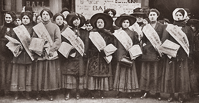 Foto av en rad kvinnor i helfigur och bakom dem anas fler kvinnor. De har banderoller över axlarna och papper i händerna. Flera har hatt eller sjal