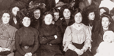 Foto av strejkande textilarbeterskor som sitter samlade. En kvinna längst till höger har ett barn i knät. I mitten sitter en kvinna med armarna i kors och ser bestämt in i kameran, bakom henne sticker en leende flicka fram huvudet. De flesta ser allvarliga ut.