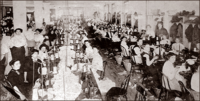 Foto av en stor sal där kvinnor sitter och jobbar med textilarbeten. Det ligger tygskräp på golven mellan borden. Längs med ena väggen anas upphängda ytterkläder.