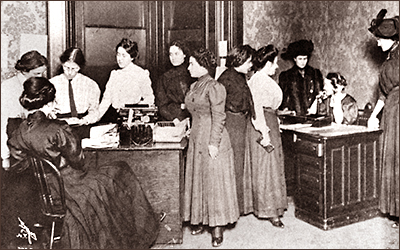 Foto av elva kvinnor i helfiogur på ett kontor. De står i grupper runt två bord och vid varje bord sitter en kvinna. Den ena tycks tala i telefon.