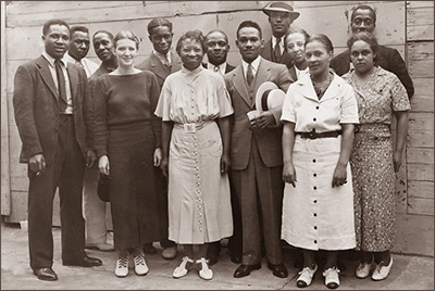 Foto av tretton personer i helfigur, stående på en gata mot en trävägg