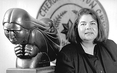 Foto av Wilma Mankiller bredvid en  staty av en Cherokeekvinna med barn på ryggen. I bakgrunden syns symbolen för Cherokee Nation på väggen.