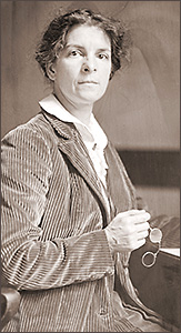 Foto av Rheta Childe Dorr 1913. Hon sitter i hanvfigur och ser in i kameran. Hon har en manchesterkavaj på sig och sina glasögon i ena handen