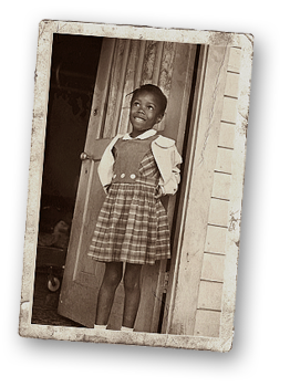 Foto av Ruby Bridges som barn. Hon står i en dörröppning och har en rutig klänning på dig samt en ljus jacka eller kofta