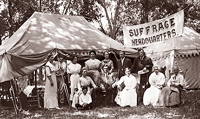 Fot av en samling kvinnor och några män framför flera stora tält, några sitter, några står. Mellan tälten hänger en banderoll med texten: Suffrage Headquarters
