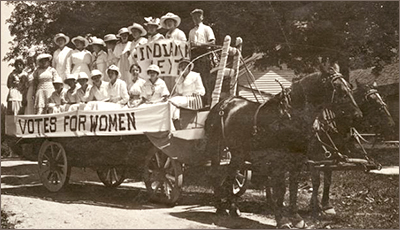 Foto av ljust klädda kvinnor på en vagn dragen av hästar. Vagnen har en banderoll med texten Votes for women