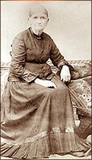 Foto av Nancy Johnson i helfigur på gamla dar, sittande iförd en lång mörk klänning