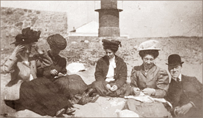 Foto av fyra kvinnor och en man som sitter på en strand med en mur bakom sig. Allla är ordentligt påklädda och har hattar på sig