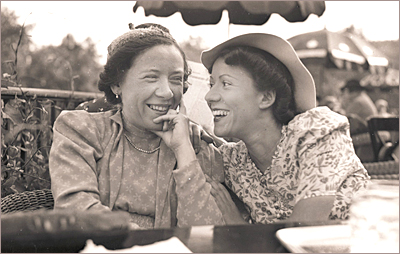 Foto av två kvinnor som sitter utomhus, kanske på ett kafé och skrattande tittar på varandra