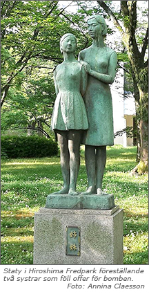 Foto av ärggrön staty av två flickor som står rakt upp och ner, den längre står lite bakom den andra och håller handen på hennes axel. I bakgrunden gräs med vita blommor, buskar och träd.