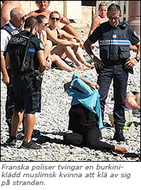 Foto av tre franska poliser som står på en stenig strand runt en sittande kvinna som håller på att klä av sig. Solande folk i bakgrunden.