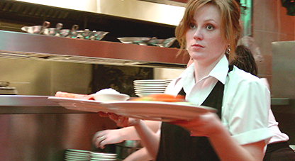 Foto av servitris med en tallrik i var hand och typiskt restaurangmiljö/kök i bakgrunden. Hons er trött ut och stirrar rakt framför sig.