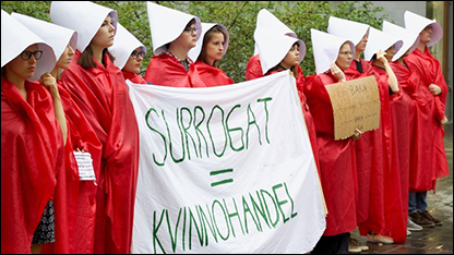 Foto av en rad kvinnor iförda kläder från tv-serien Handmaid's Tale. De håller en banderoll med texten surrogat = kvinnohandel