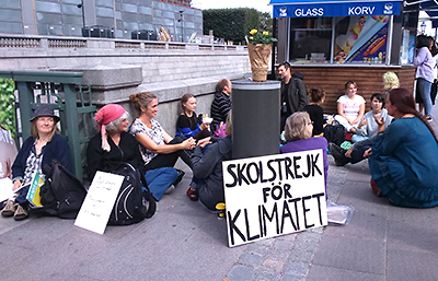 Foto av ett gäng strejkande sittande på marken med Greta mitt i. Närmast i bild står ett plakat med texten "Skolstrejk för klimatet". I bakgrunden syns en glass- och korvkiosk