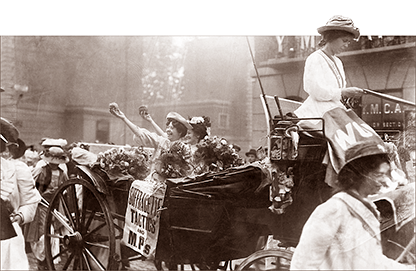 Foto av två kvinnor som vinkar med blomsterkvastar i armarna i en vagn som dras av häst utom bild, men kvinnan som sitter på kuskbocken syns. Runt om går glada och hurrande kvinnor