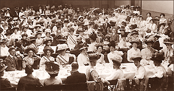 Foto av massor av kvinnor, de flesta iförda stora hattar, och några män i en stor sal runt flera avlånga bord