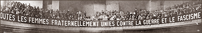 Avlångt foto av en banderoll som hänger på en balkong (öppen övervåning i en lokal) med massor av kvinnor runt om. På banderollen står: Toutes les femmes fraternellement unies contre la guerre et le fascisme