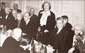 Foto av en grupp män och kvinnor som sitter längs med ett bord. Mitt i bilden står en kvinna upp och ser in i kameran