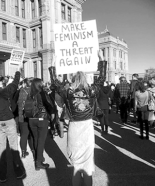 Foto av demonstrationståg med folk sedda bakifrån. I centrum på bilden går en kvinna som håller upp ett plakat som det står "Make feminism a threat again" på.