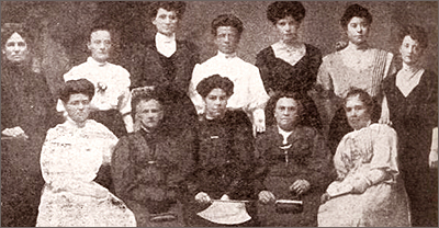 Foto av tolv kvinnor i gammaldags klänningar, fem som sitter och sju som står bakom dem. Alla ser in i kameran
