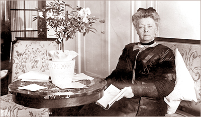 Foto av Bertha von Suttner sittande i en soffa vid ett runt bord med blommor på. Hon håller en bok i händerna och ser in i kameran