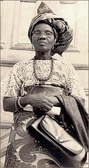 Foto i nästan helfigur av Funmilayo som håller i en handväska. Hon har sjal på huvudet, glasögon och ett halsband